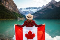 woman wearing red and black checkered dress shirt and beige fedora hat holding canada flag looking at lake. You can drive in alberta for 90 days with out of province license.